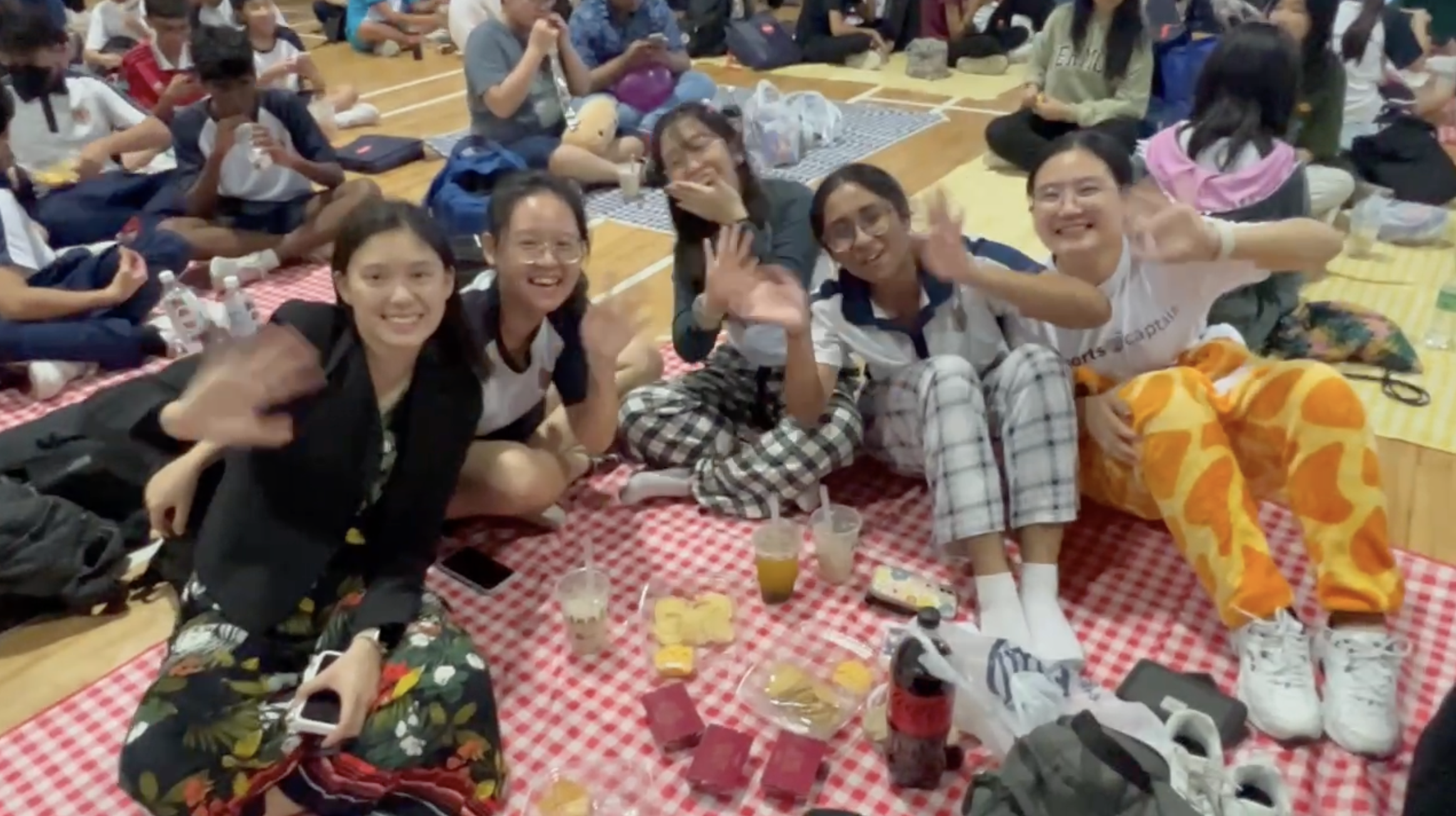 Five friends sit on a red checkered mat, smiling and waving, with snacks and drinks on a gym floor.
